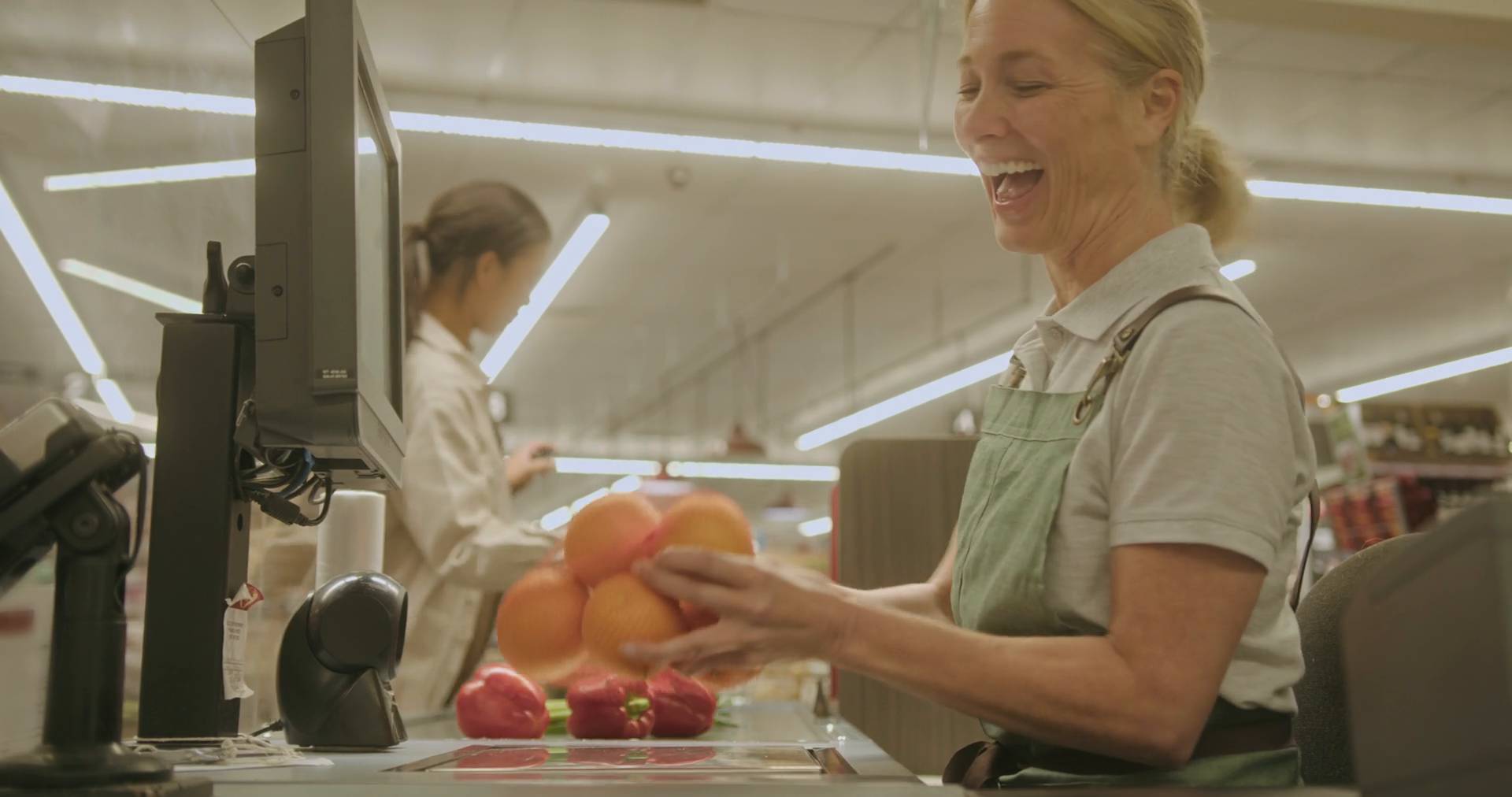 Cajera de supermercado sonriendo mientras pasa frutas y verduras por la caja.